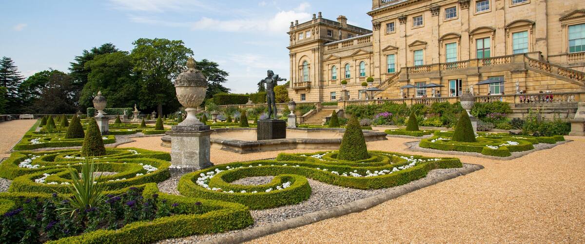 A grand historic building with formal gardens, including trimmed hedges and statues.