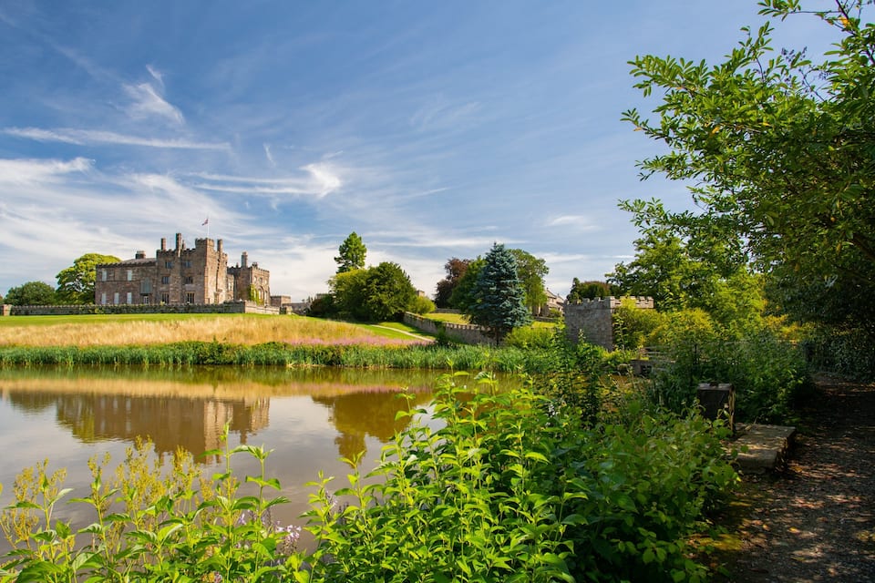 A castle by a calm lake surrounded by lush greenery and a clear sky.