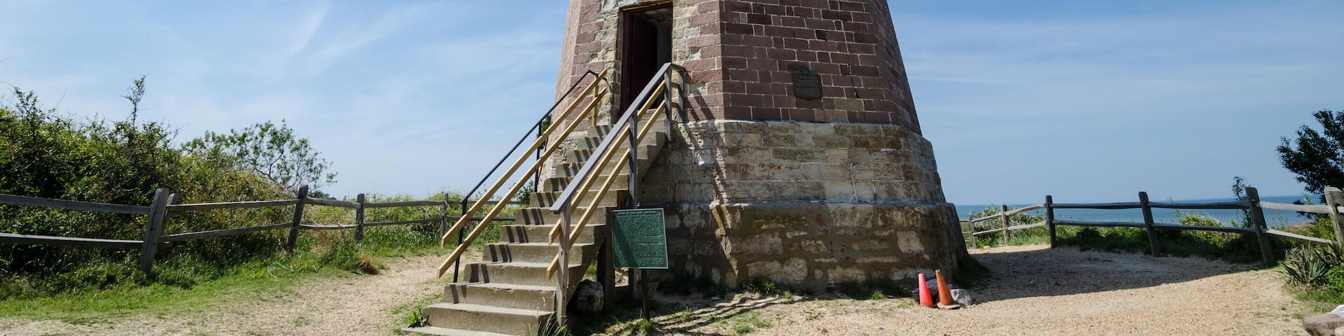 A tall brick tower with a staircase leading up to a door, surrounded by a wooden fence and a clear sky.