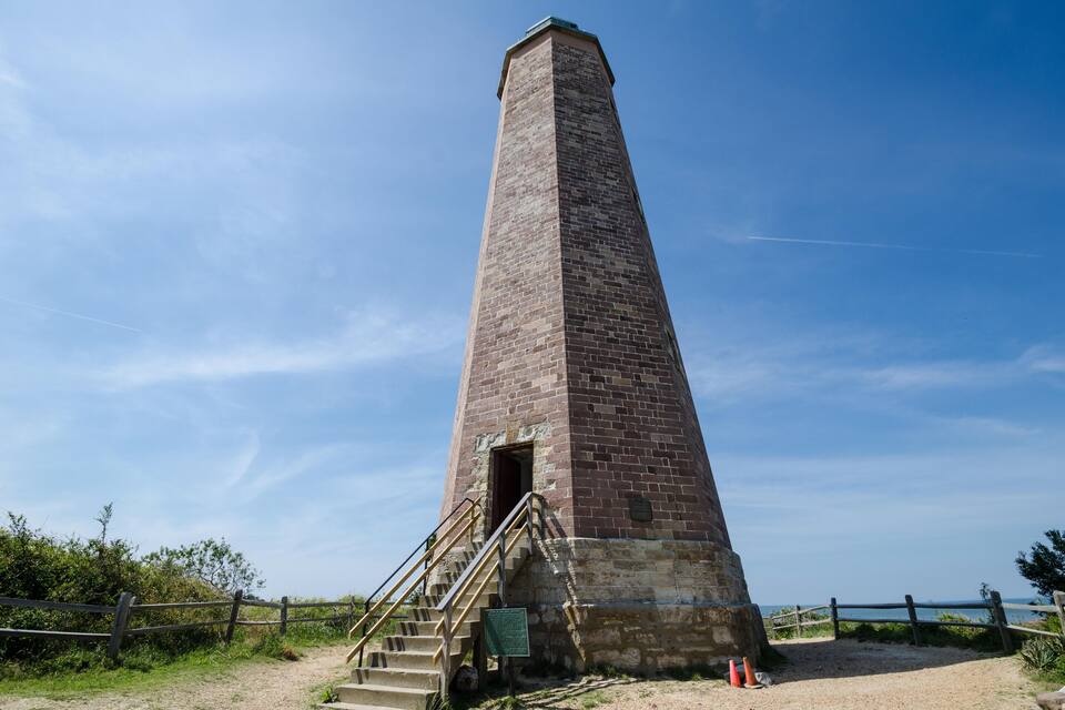 Una alta torre de ladrillo con una escalera que sube a una puerta, rodeada por una valla de madera y un cielo despejado.