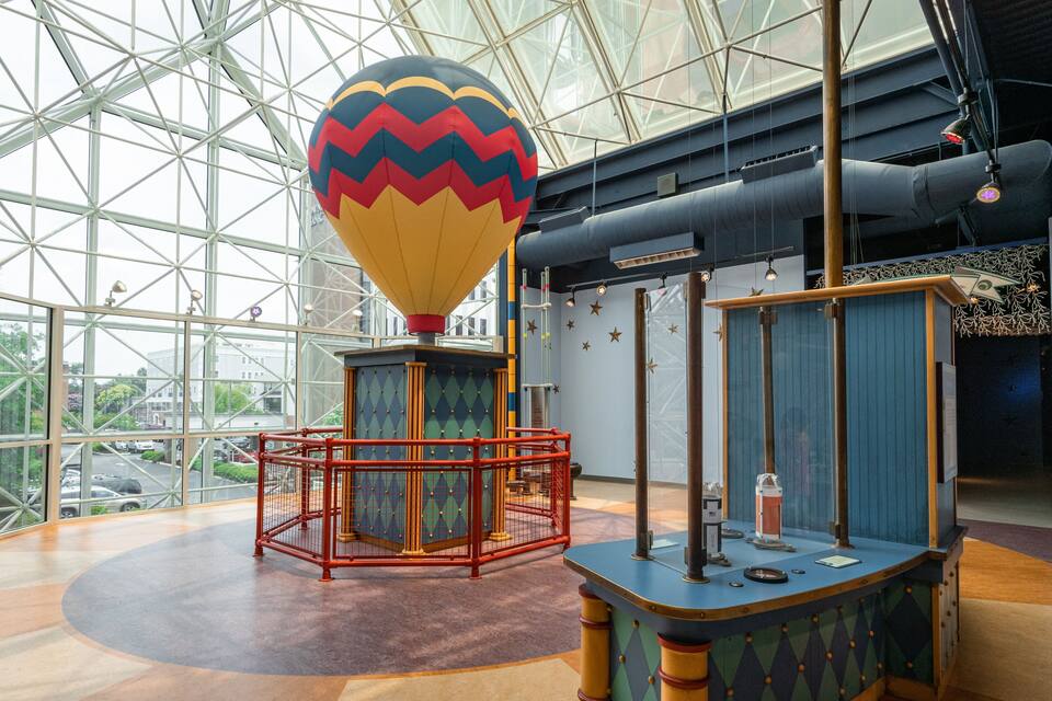 A large glass-enclosed exhibit with a colorful hot air balloon, a red metal play structure, and a blue table with scientific equipment.
