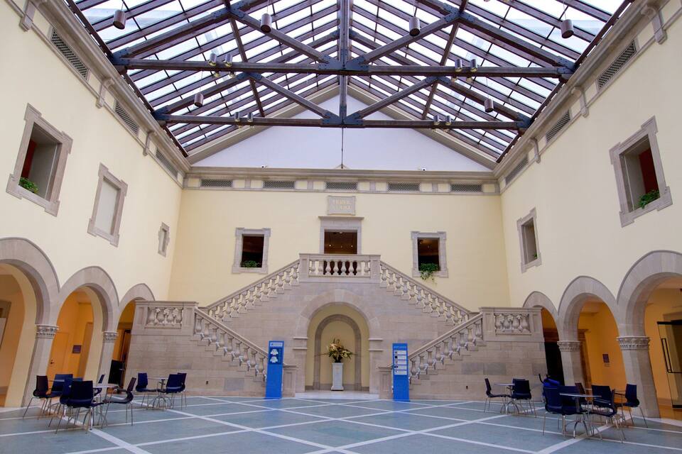 A spacious atrium with a high ceiling and a glass roof, featuring a central staircase with ornate balustrades and a floral arrangement at the top.