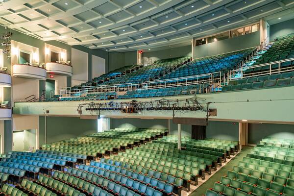 A large indoor auditorium with multiple levels of seating, green chairs, and a stage area with technical equipment.