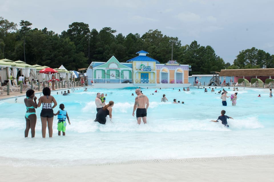 Un parque acuático con una piscina grande, zonas de playa y varios edificios, incluido un puesto de socorristas.