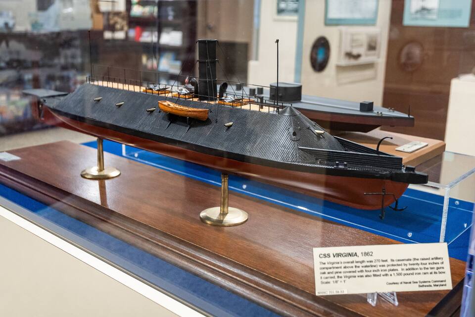 A model of the CSS Virginia, 1862, a Confederate ironclad warship, displayed on a wooden stand with a plaque.