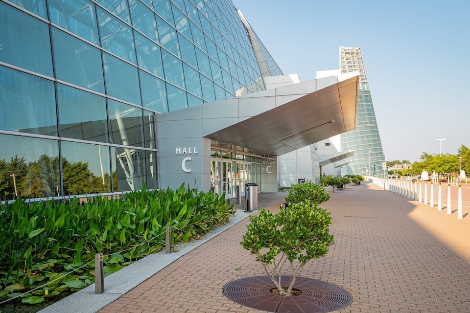 A modern building with reflective glass windows, a sign reading "HALL C", and a paved walkway with greenery and bollards.