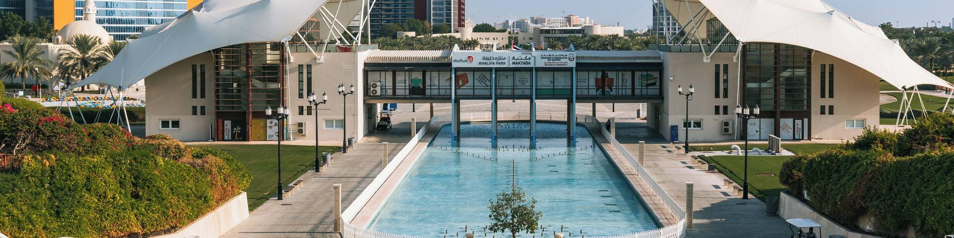 A modern building with a pool in front, surrounded by greenery and palm trees.