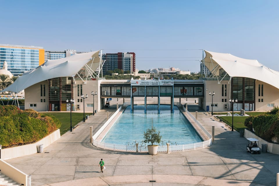 A modern building with a pool in front, surrounded by greenery and palm trees.