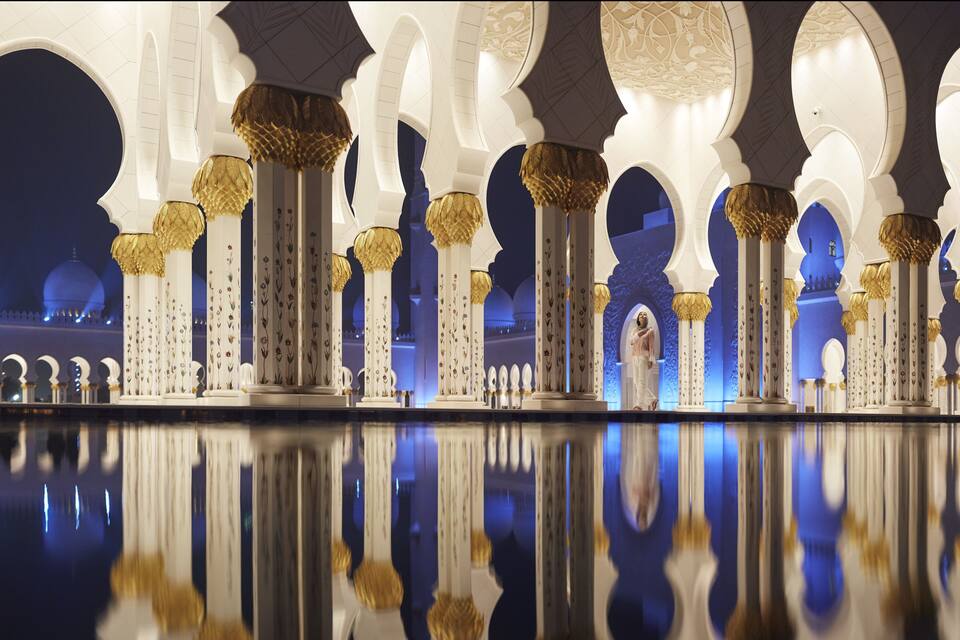 A series of white columns with golden tops and floral patterns, reflecting on a polished floor.