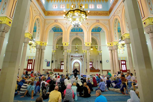 Une vaste salle richement décorée, dotée de hauts plafonds, de lustres et où une foule de personnes est assise sur le sol.