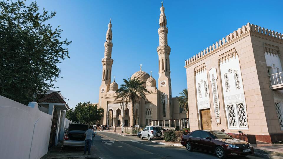 A mosque with two minarets and a large dome, surrounded by a street with cars and pedestrians.