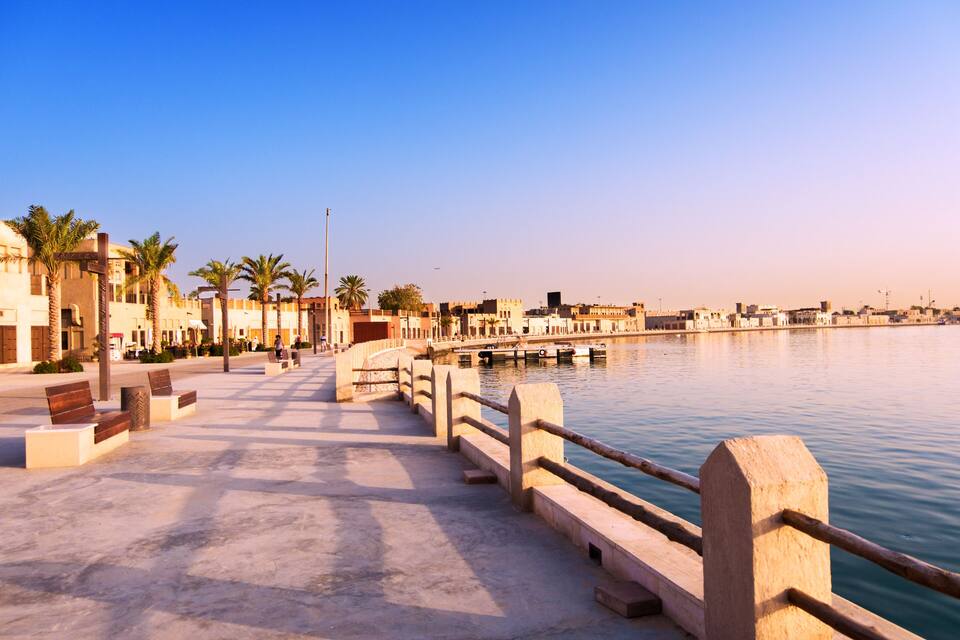Um calçadão à beira-mar com palmeiras, bancos e uma ponte.