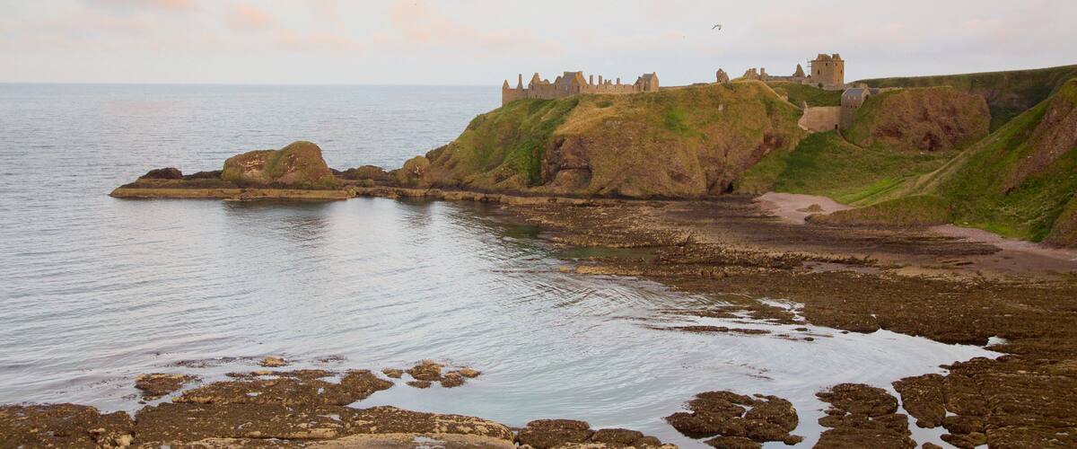 A coastal landscape with a castle on a grassy cliff overlooking the sea.