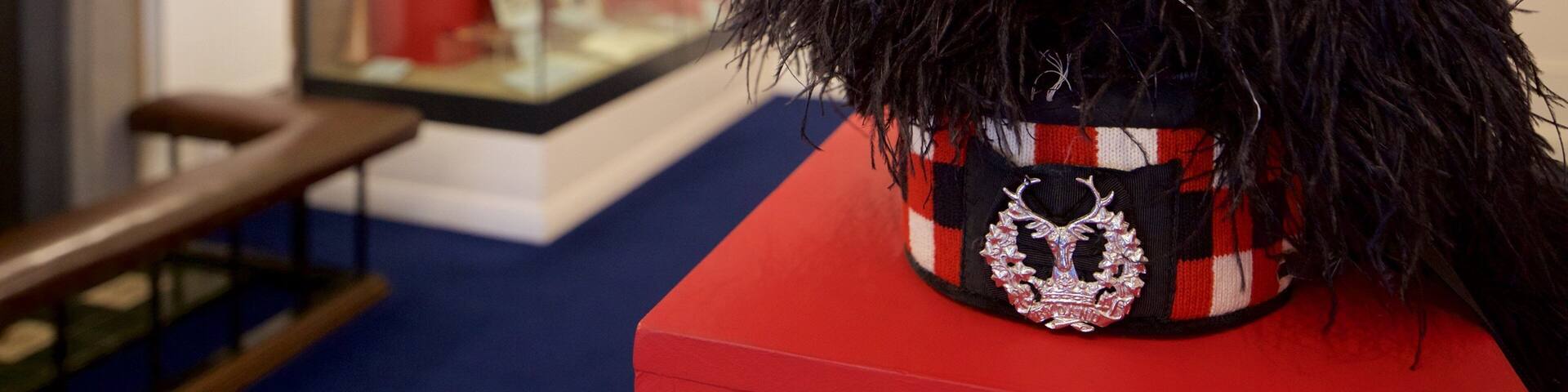 A display of a black, textured hat with a decorative emblem on a red and black striped base, set against a white wall with a blue carpet.