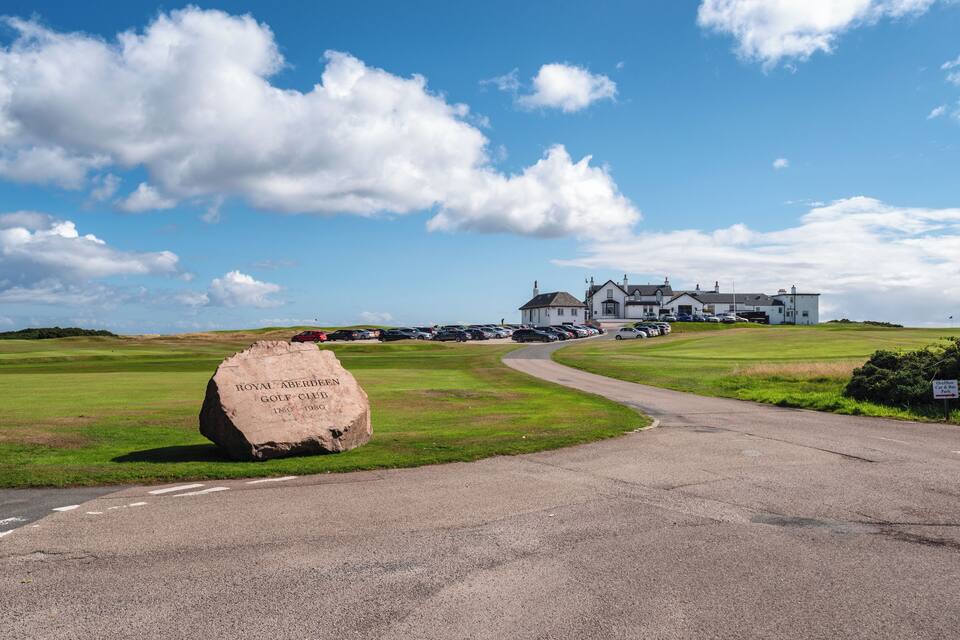 A golf course with a large rock inscribed with "Royal Aberdeen Golf Club 1868-1968" and a clubhouse in the background.