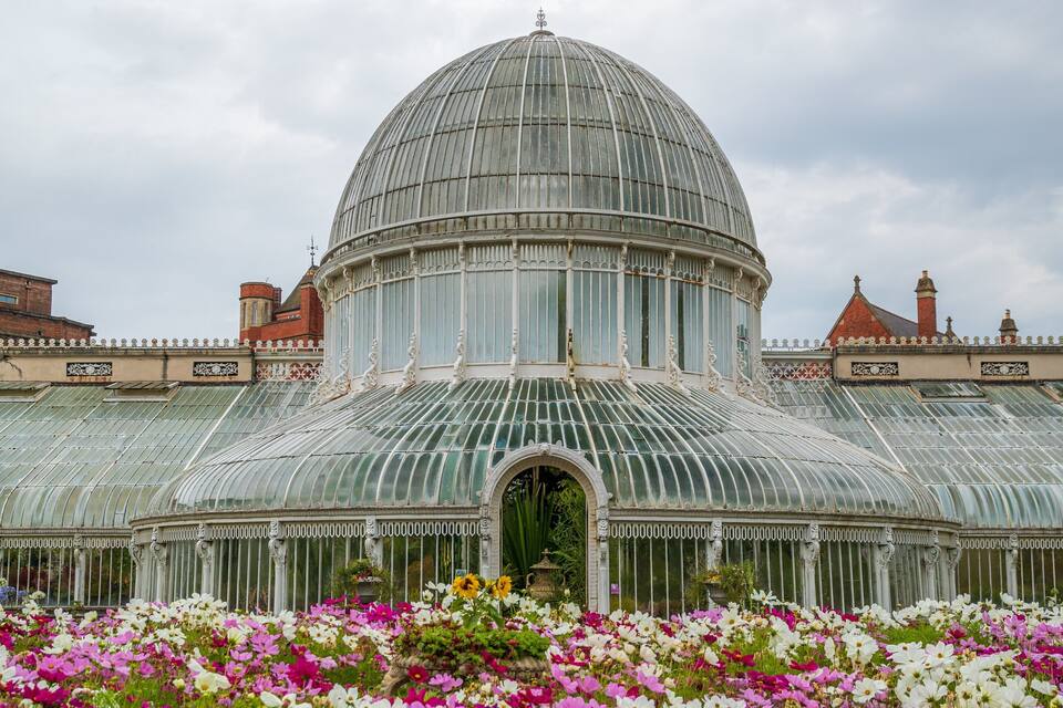 A large glass-domed greenhouse with a metal roof, surrounded by colorful flowers.