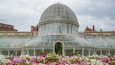 A large glass-domed greenhouse with a metal roof, surrounded by colorful flowers.