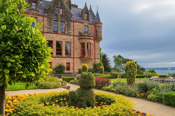 A historic stone building with red windows and a turret, surrounded by well-manicured gardens and a lake in the background.