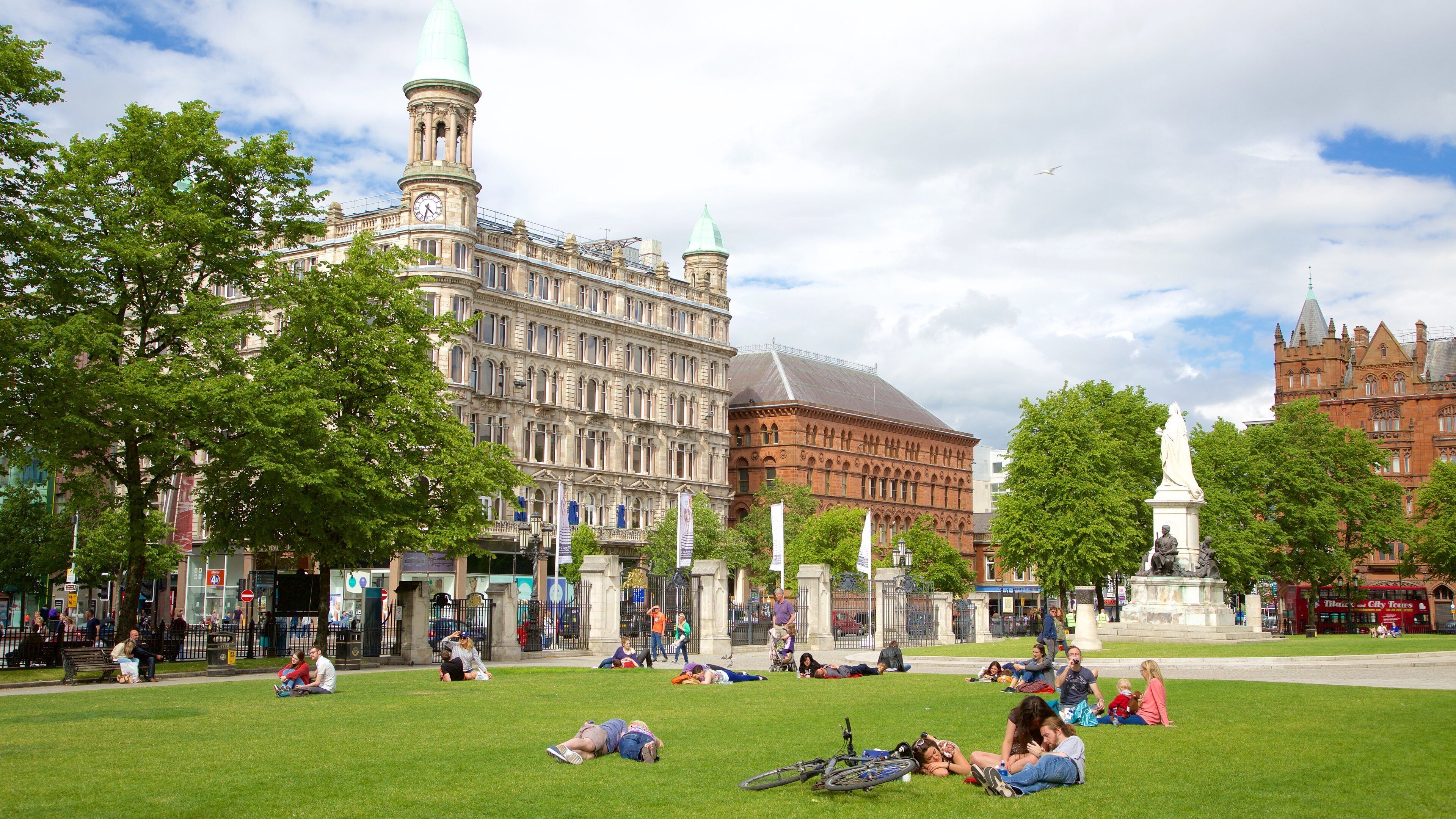 A park with people relaxing on the grass, surrounded by historic buildings and a statue.