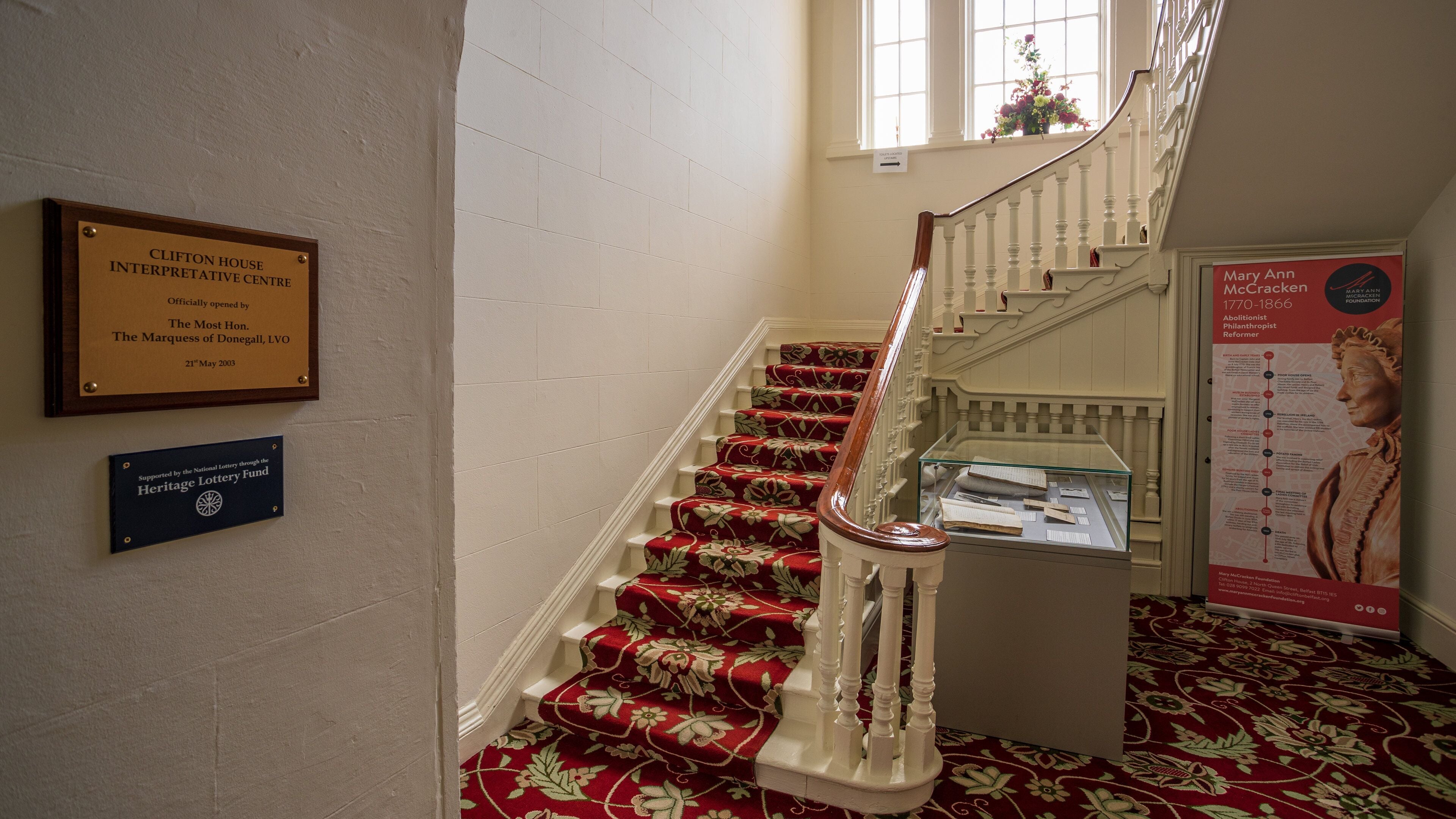 Un escalier dont les marches sont recouvertes d’une moquette rouge, une plaque commémorative sur le mur et une vitrine contenant des documents.