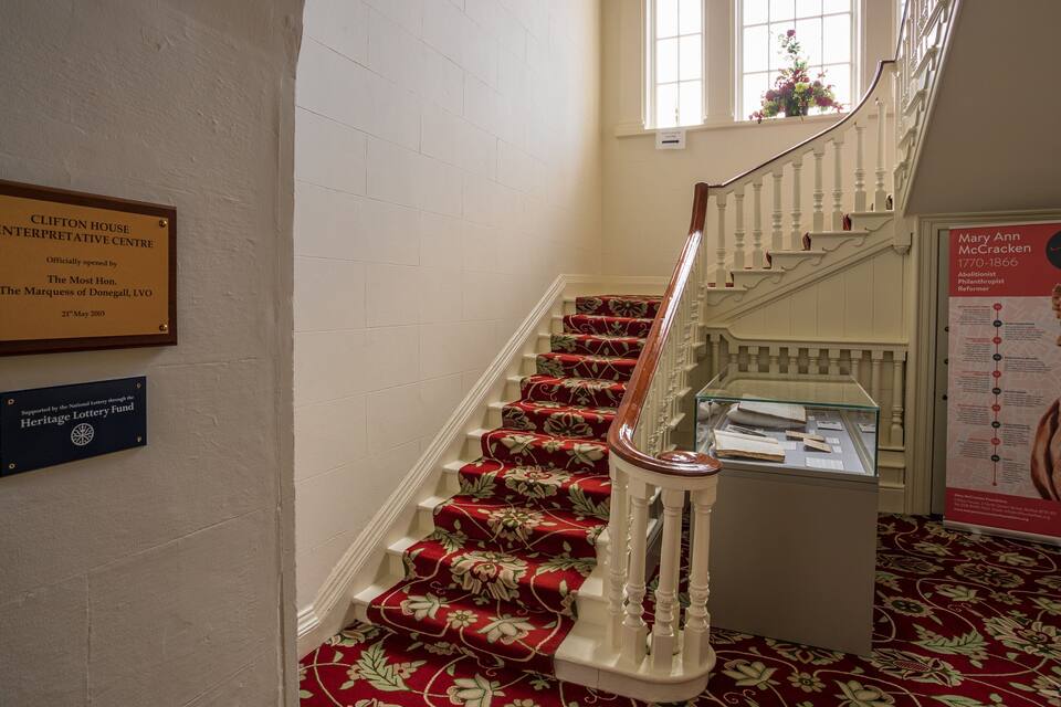 Un escalier dont les marches sont recouvertes d’une moquette rouge, une plaque commémorative sur le mur et une vitrine contenant des documents.