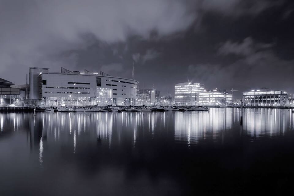 Uma marina noturna com barcos atracados, um edifício moderno com vários andares e um hotel bem iluminado ao fundo.