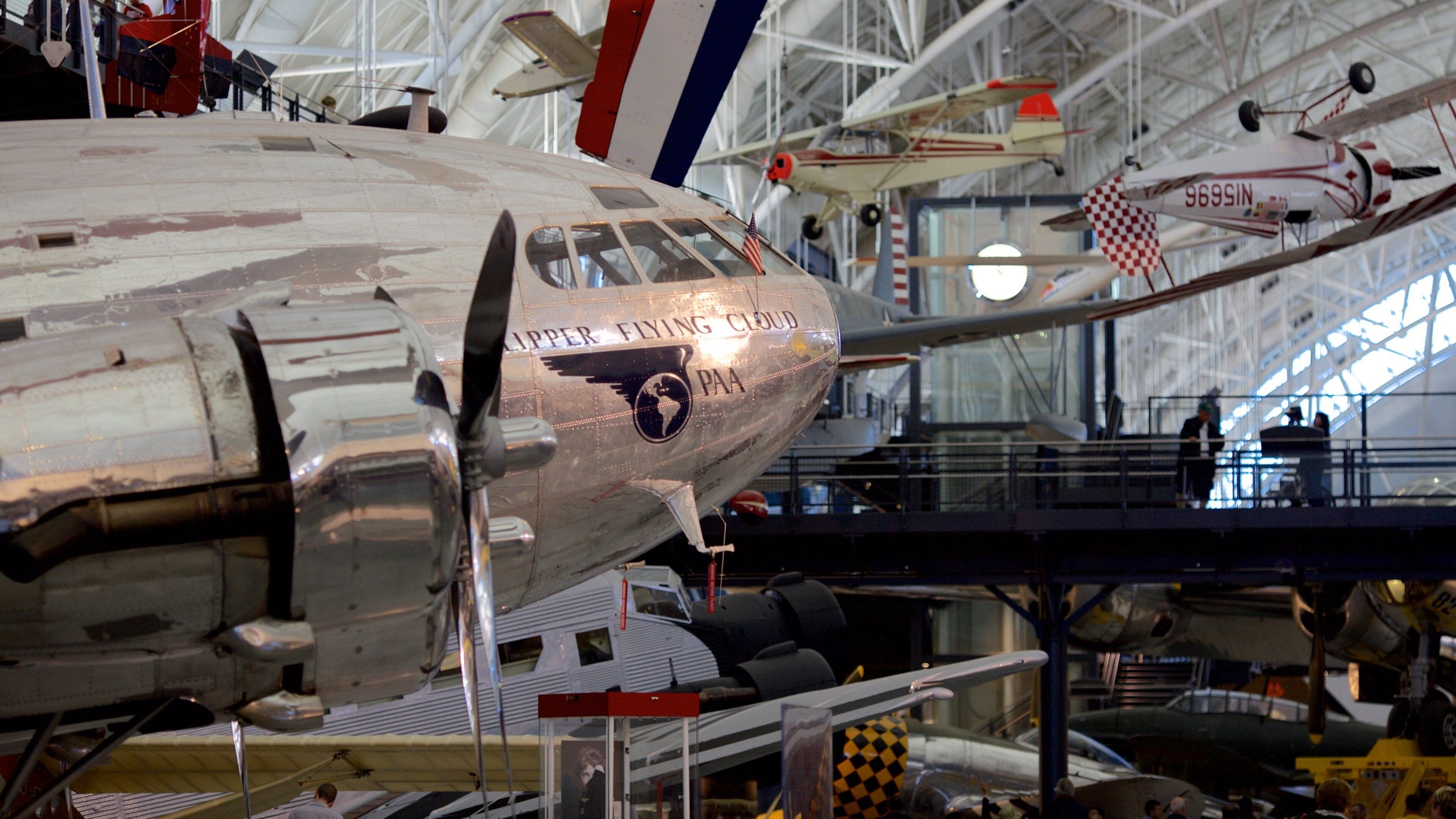 Un avión antiguo con la inscripción "UPPER FLYING CLOUD" y "PAA" en su fuselaje, exhibido en un museo cubierto junto a otras aeronaves.