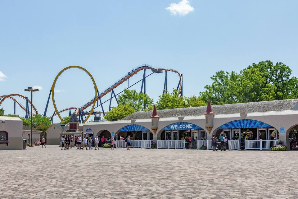 Des montagnes russes dans un parc d’attractions, avec une pancarte indiquant « Bienvenue » et une foule de personnes.
