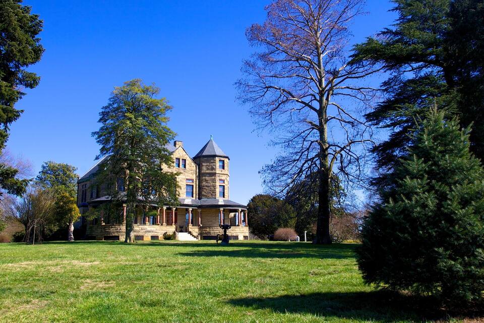 A large stone mansion with a turret, surrounded by a well-maintained lawn and tall trees.