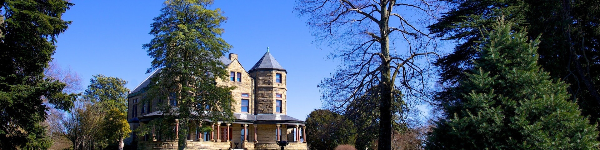 Uma grande mansão de pedra com uma torre, cercada por um gramado bem cuidado e árvores altas.