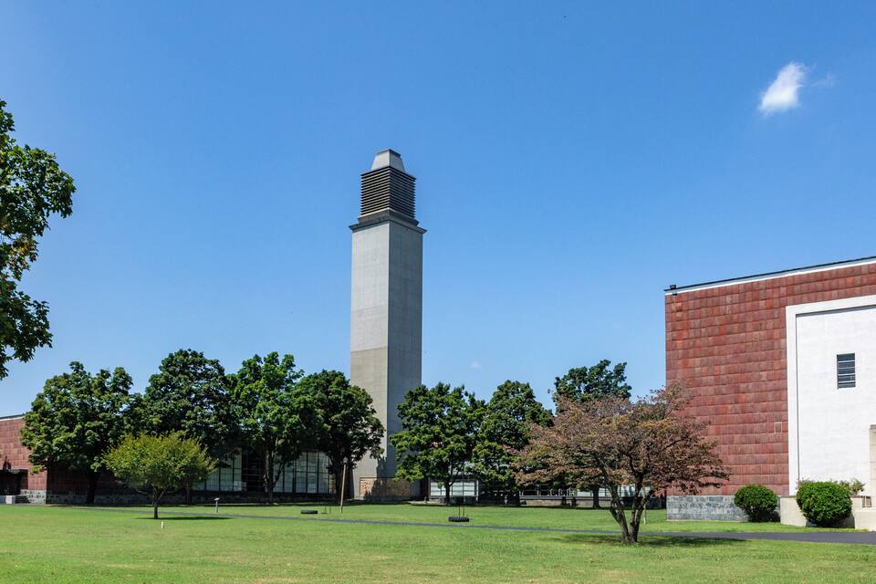 A tall monument with a square base and a pointed top, surrounded by a grassy area with trees and a red brick building.