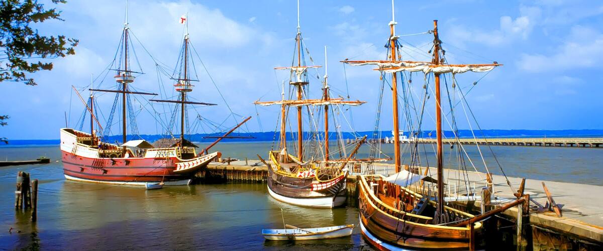 Three large sailing ships docked at a pier with a clear blue sky and calm sea in the background.