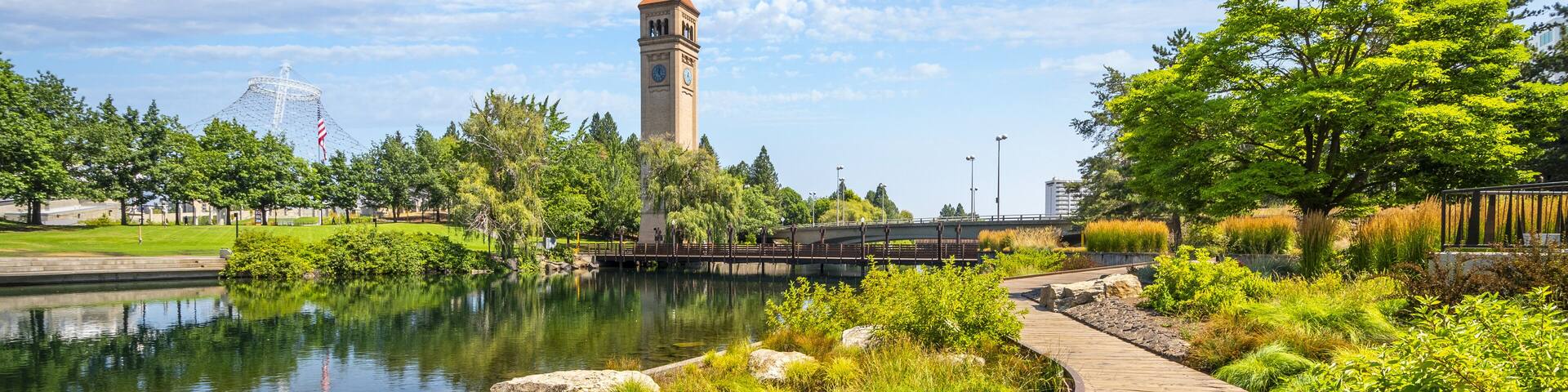 A wooden boardwalk, a tower, a body of water, and a park with trees and grass.