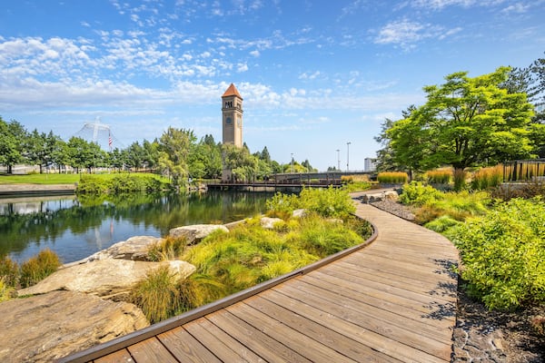 Ein hölzerner Steg, ein Turm, ein Gewässer und ein Park mit Bäumen und Gras.