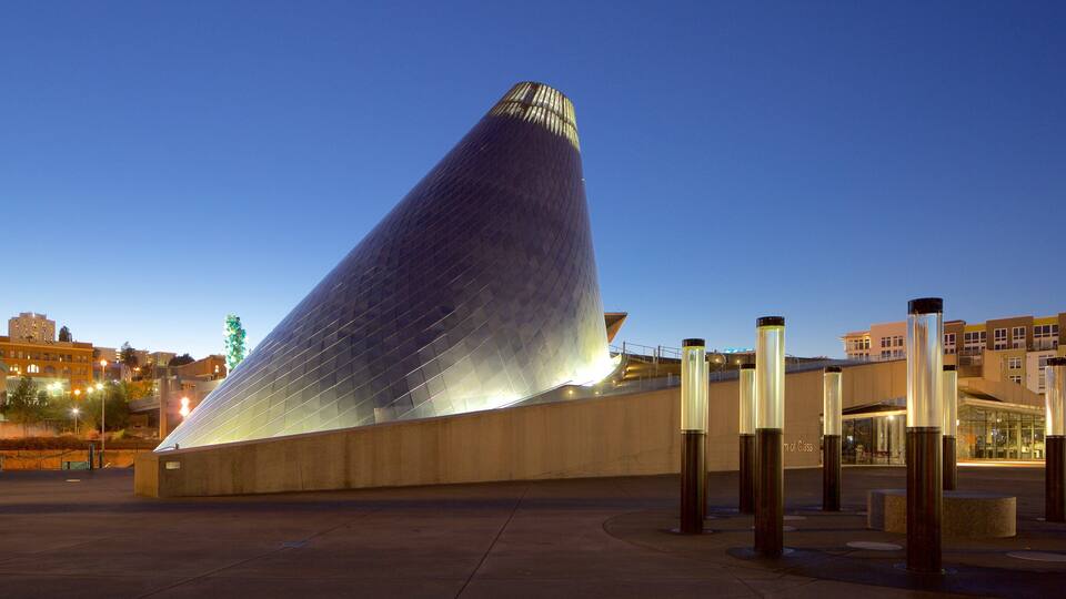 A modern architectural structure with a curved, metallic surface and a pointed top, surrounded by a plaza with bollards and buildings in the background.