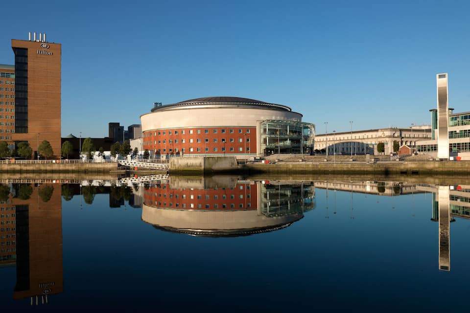 A circular building with a glass facade, a Hilton hotel, and a modern skyscraper in the background.