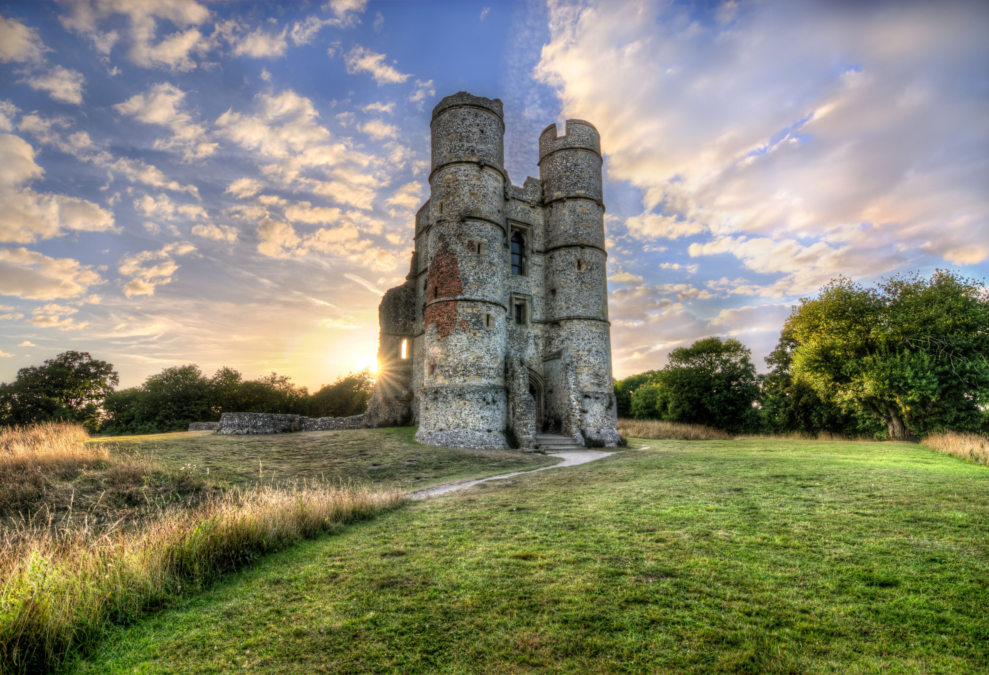A historic stone castle with two prominent towers, surrounded by a grassy field and trees, under a partly cloudy sky.