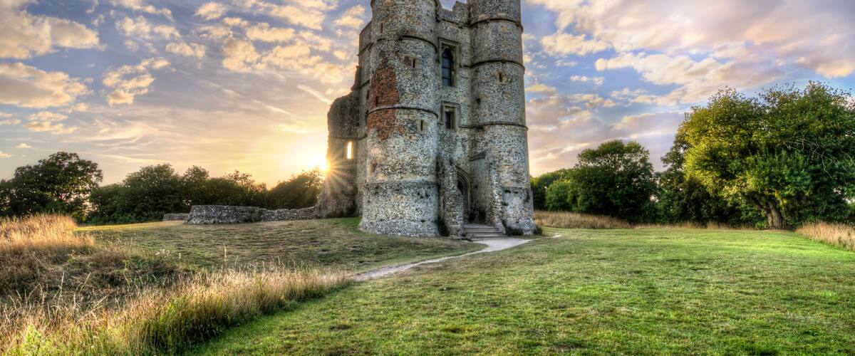 A historic stone castle with two prominent towers, surrounded by a grassy field and trees, under a partly cloudy sky.