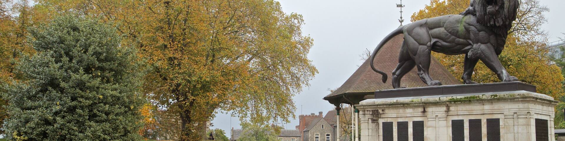 A park with a lion statue, green grass, and autumn trees.