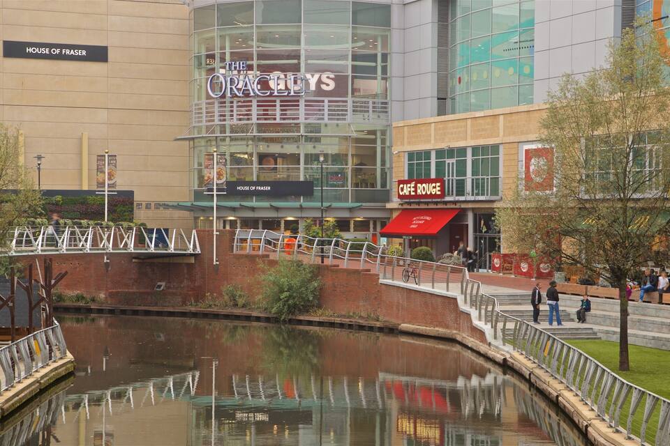 A riverside walkway with a bridge, cafes, and a modern building with a glass facade.