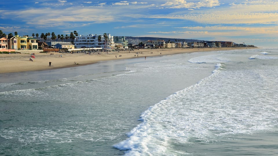 A beach with waves, a sandy shore, and buildings in the background.