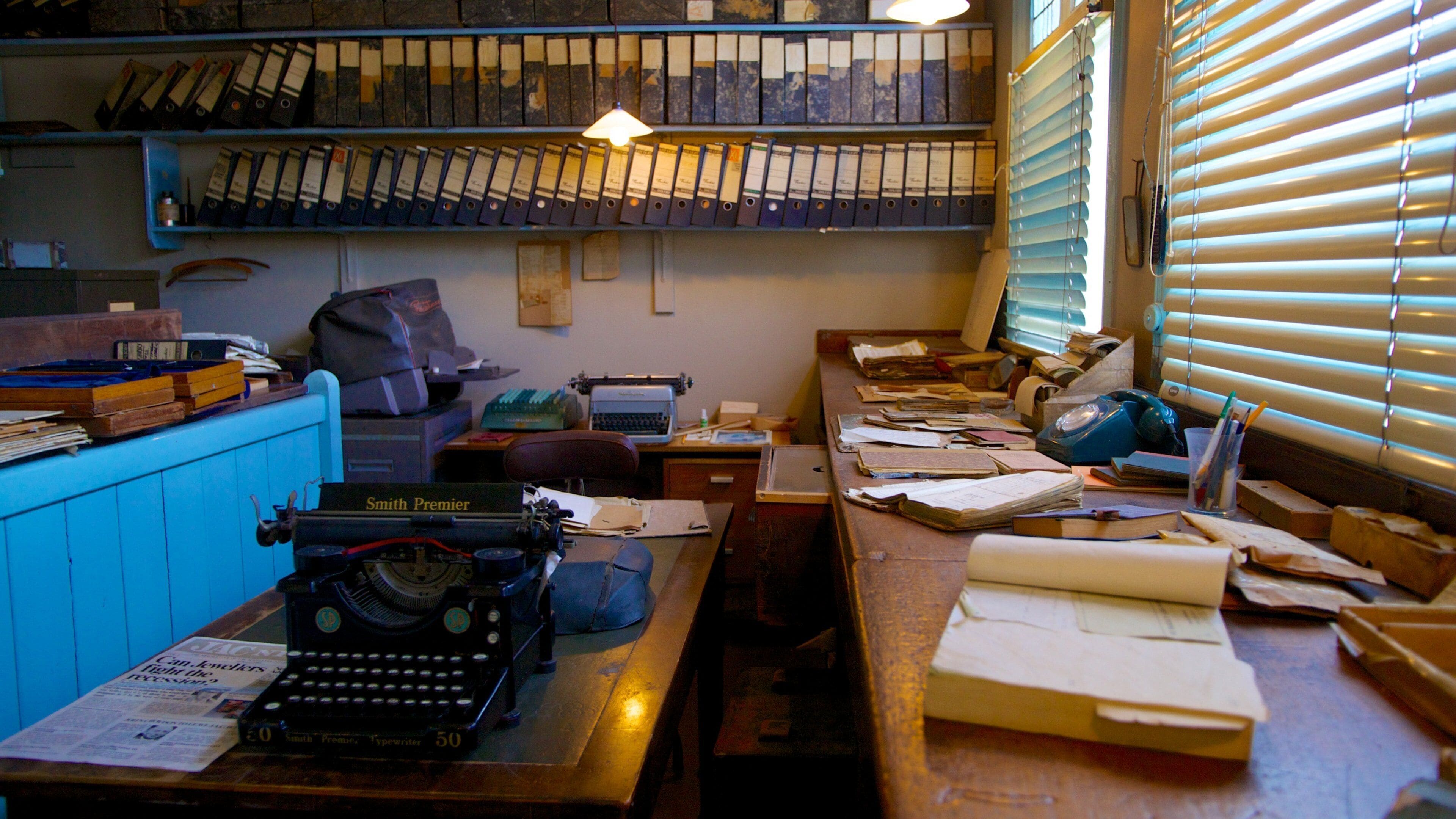 A cluttered office with a typewriter, shelves of binders, and a desk with papers and a lamp.