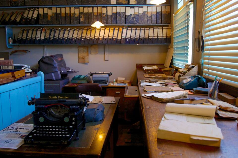 A cluttered office with a typewriter, shelves of binders, and a desk with papers and a lamp.