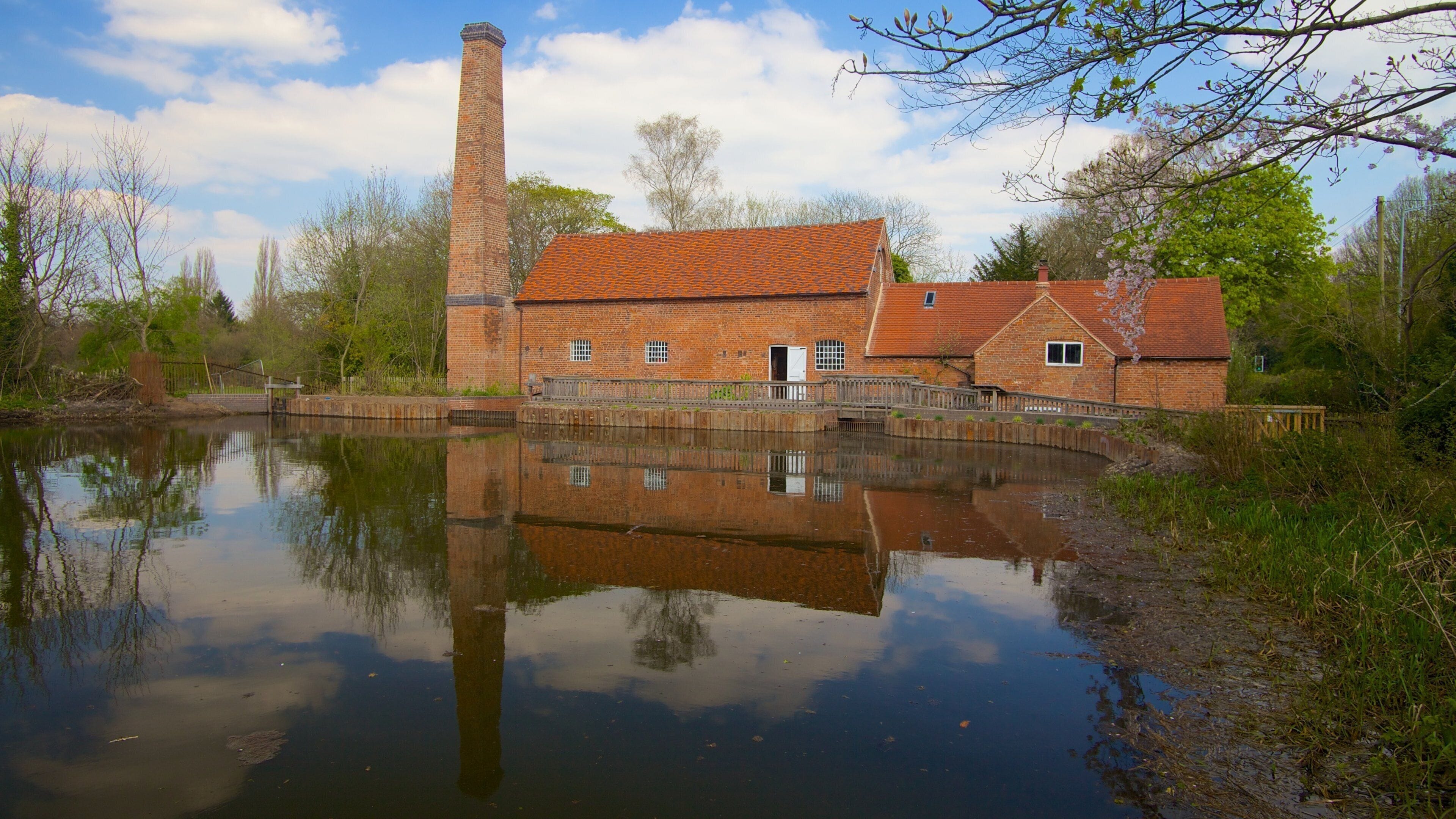 A brick building with a chimney, a pond reflecting the structure, and trees in the background.