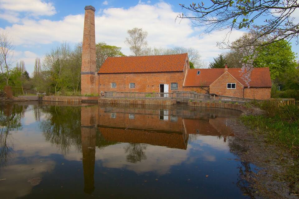 A brick building with a chimney, a pond reflecting the structure, and trees in the background.
