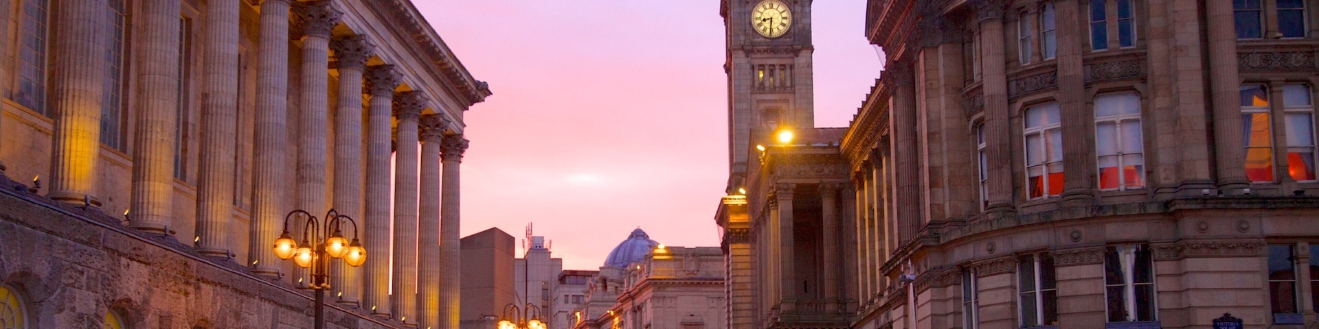 Uma rua histórica com arquitetura clássica, uma imponente torre do relógio e luminárias iluminadas.
