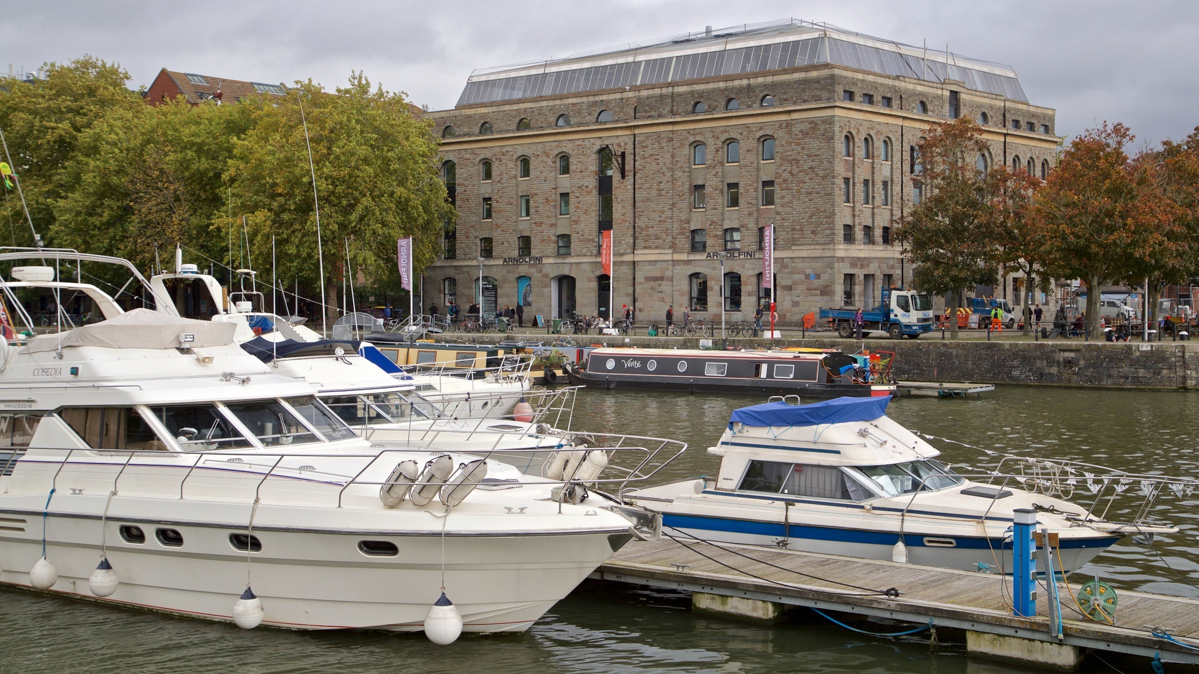 En lystbådehavn med flere både i havnen, en stor bygning med spændt buer og et bybillede i baggrunden.