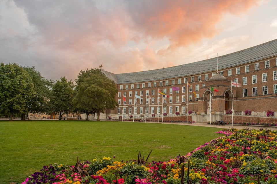 A large brick building with multiple flags, a green lawn, and colorful flower beds.