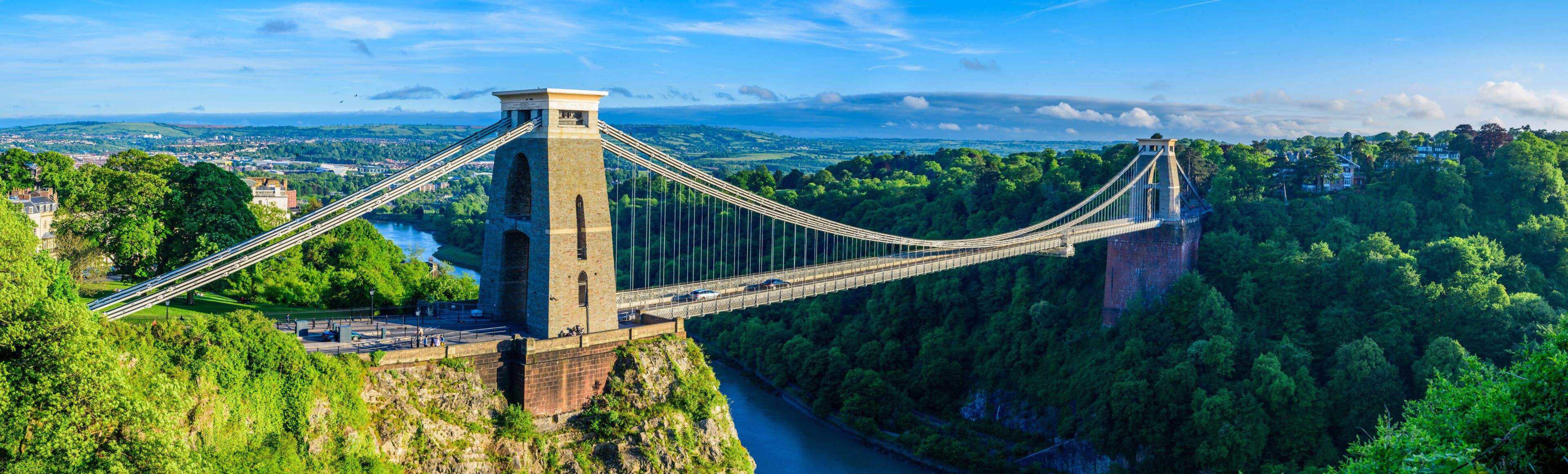A suspension bridge spanning a deep gorge, surrounded by lush greenery and a distant town.