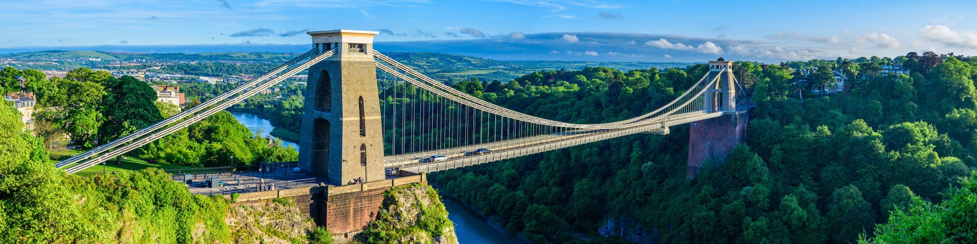 A suspension bridge spanning a deep gorge, surrounded by lush greenery and a distant town.